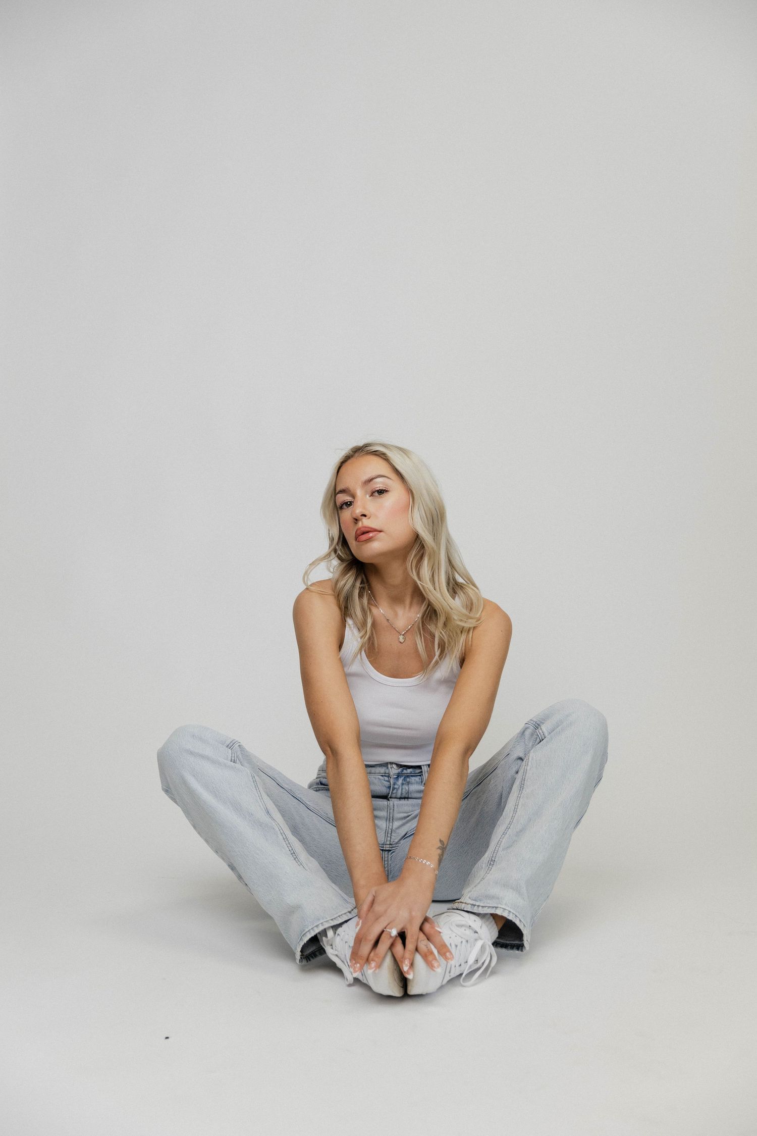 A person in light denim jeans and a white tank top sits in various relaxed poses against a white background.