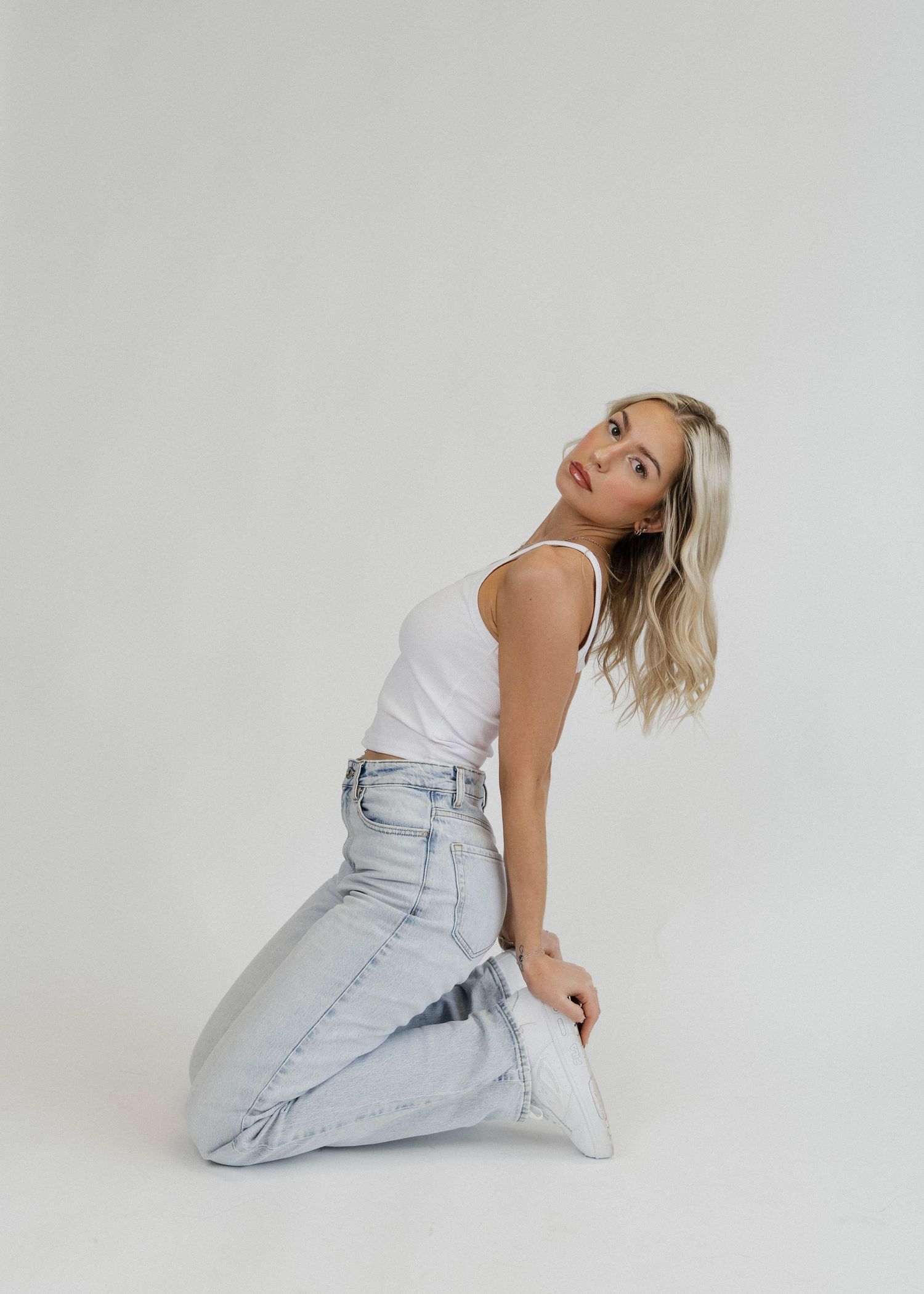 Woman in white tank top and light wash jeans poses on white background in minimalist fashion photo shoot.
