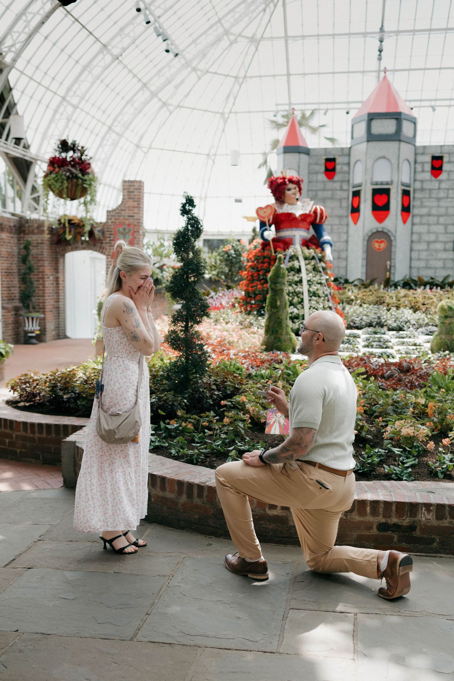 A man proposes on one knee to a woman in a floral conservatory with castle decorations and topiary sculptures.