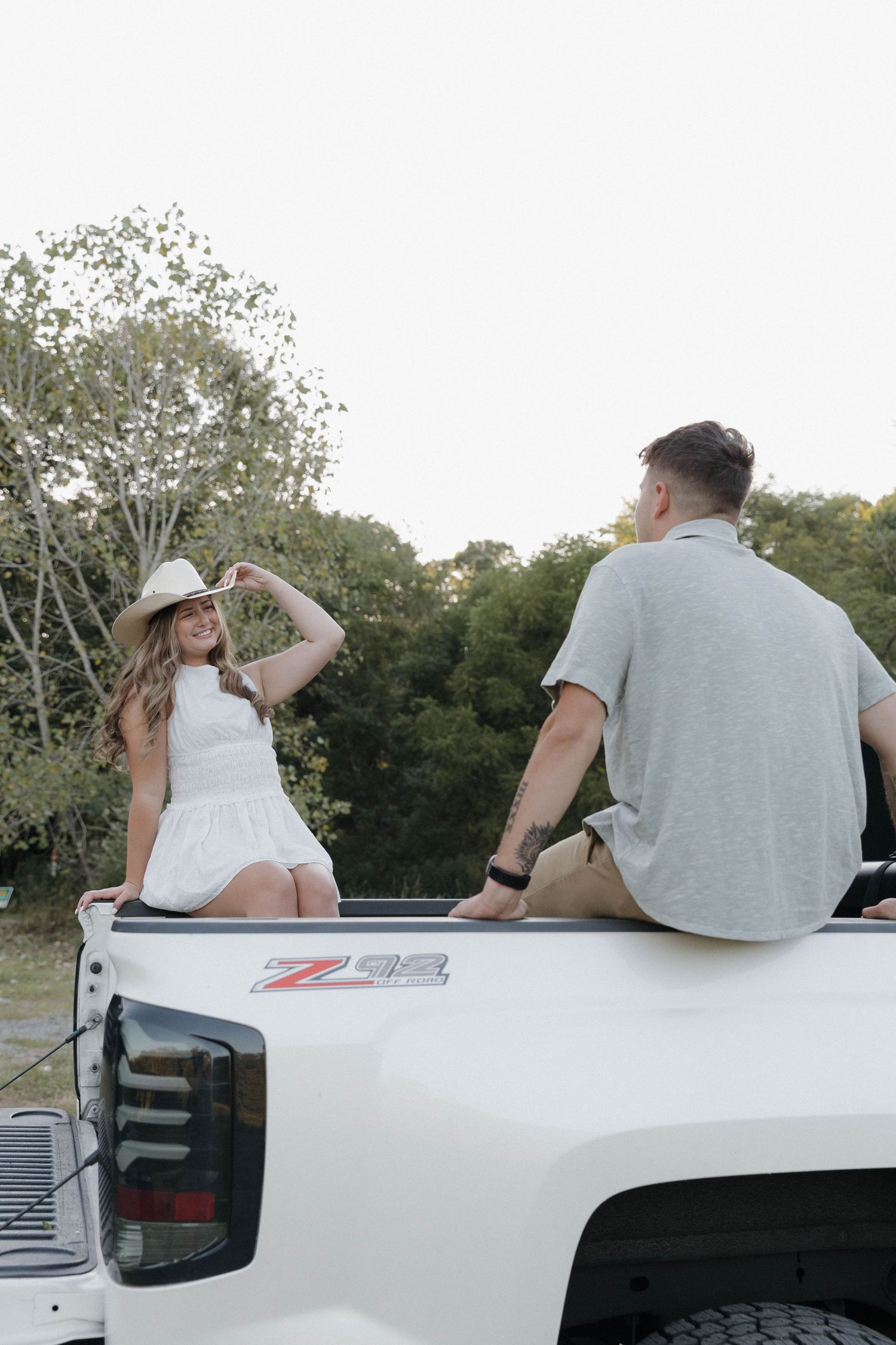 Two people sitting casually in the bed of a white pickup truck on a sunny day surrounded by trees.