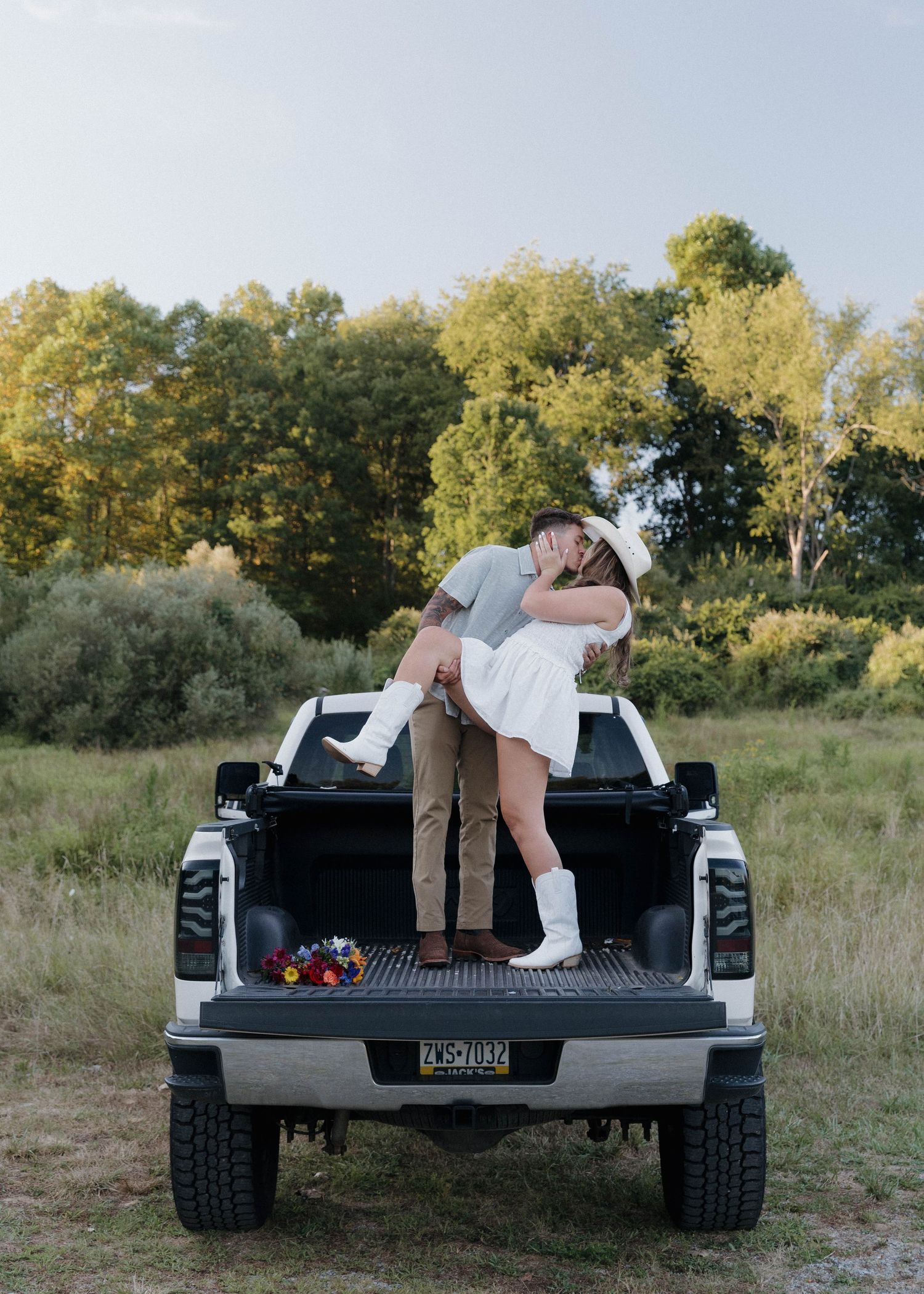 A couple embraces on the back of a black pickup truck in a rustic outdoor setting with green trees in the background.