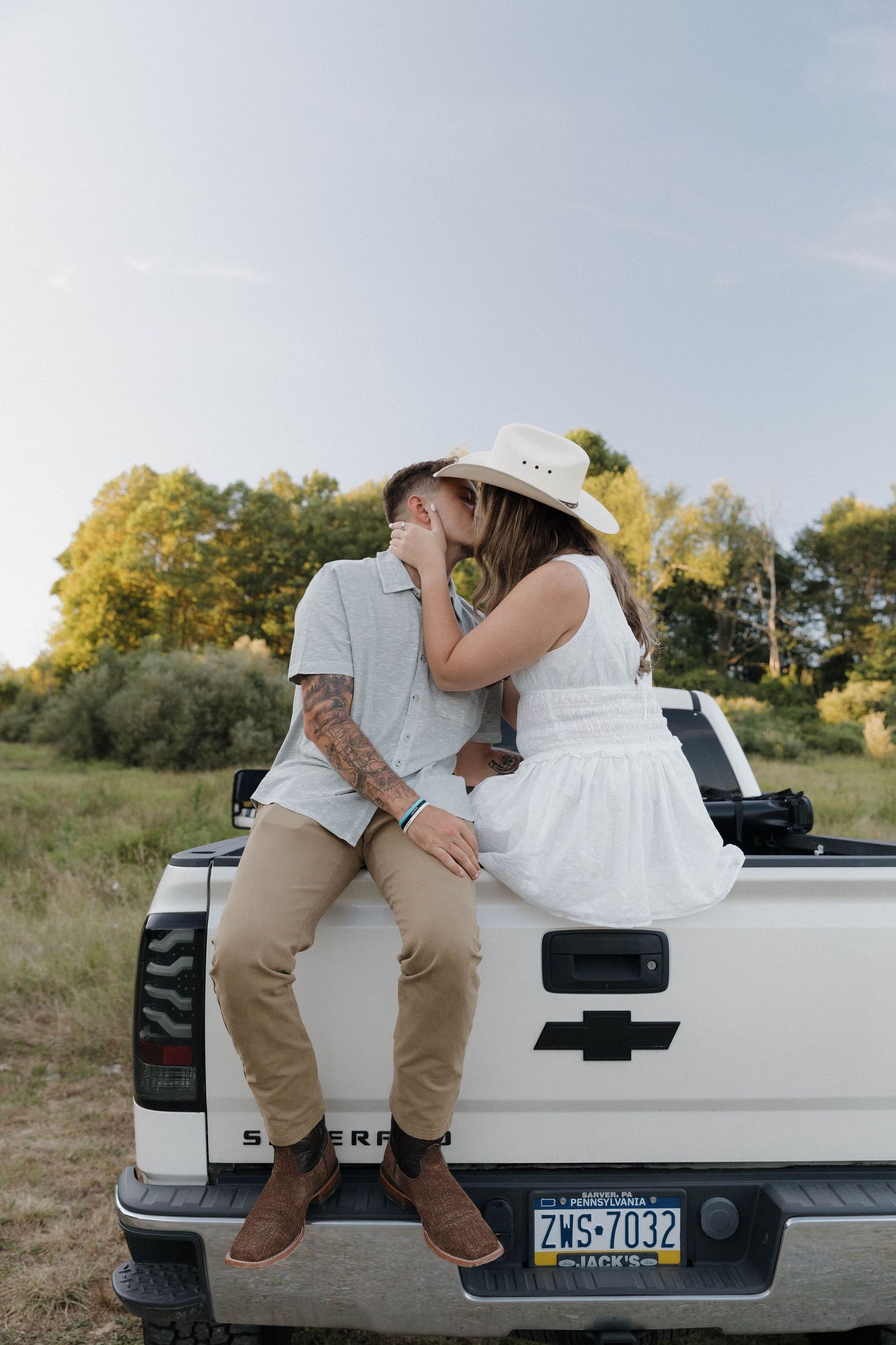 A romantic couple embraces while sitting on the tailgate of a white Chevrolet pickup truck in a rural field setting.