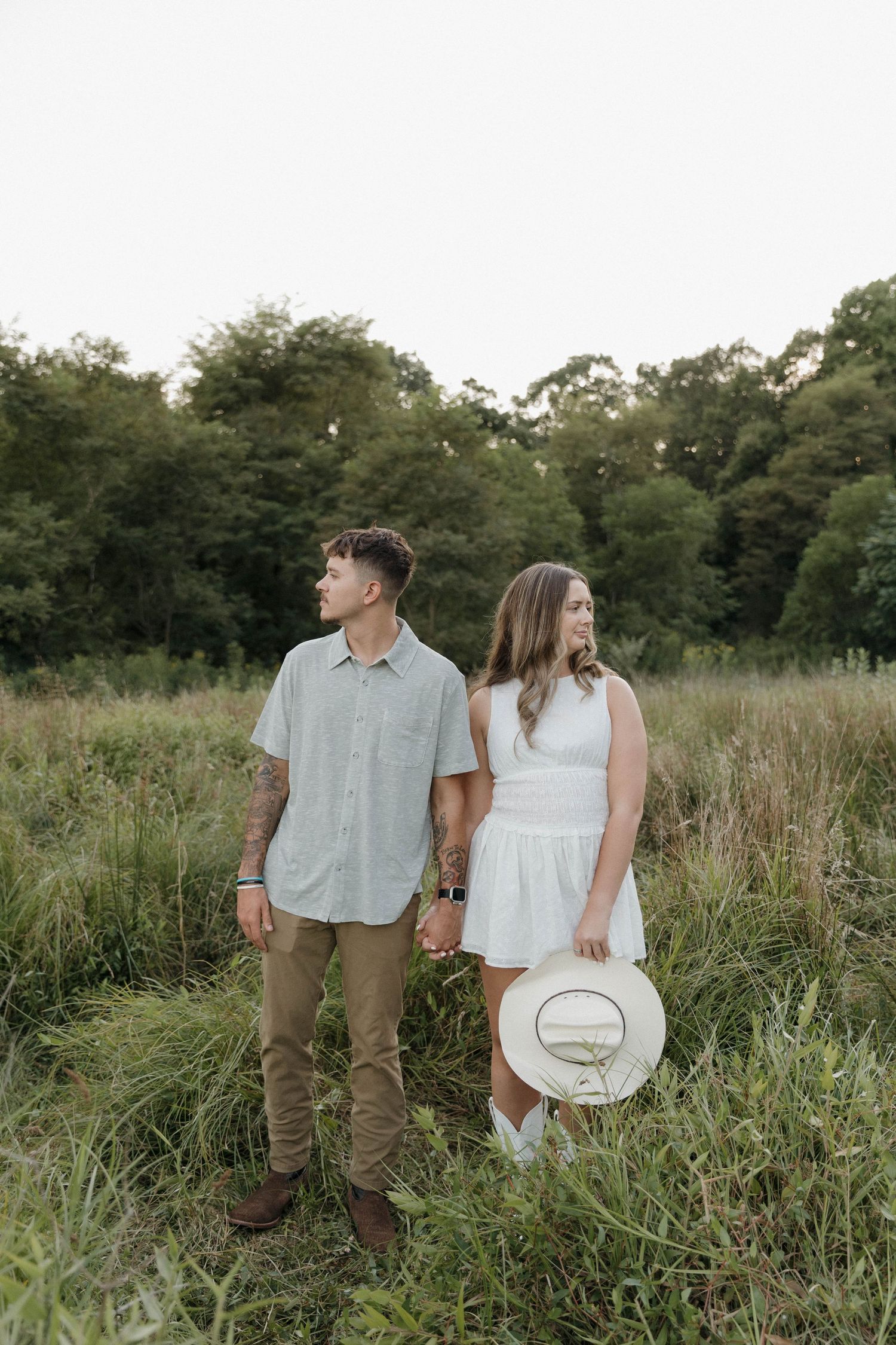 A couple stands together in a grassy meadow at sunset wearing casual summer attire with a straw hat.