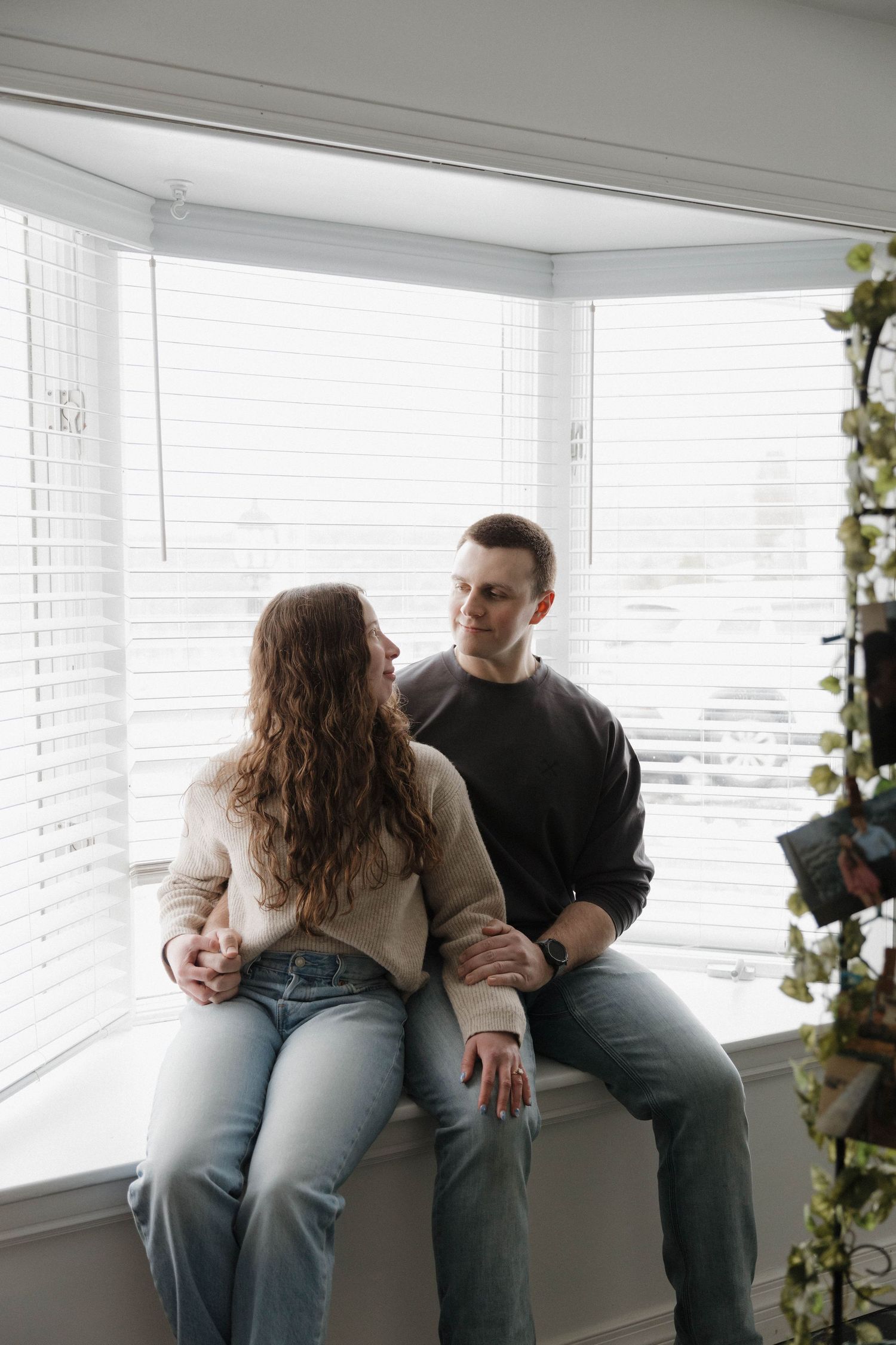 A couple sits together on a window ledge in a bright room with natural lighting.