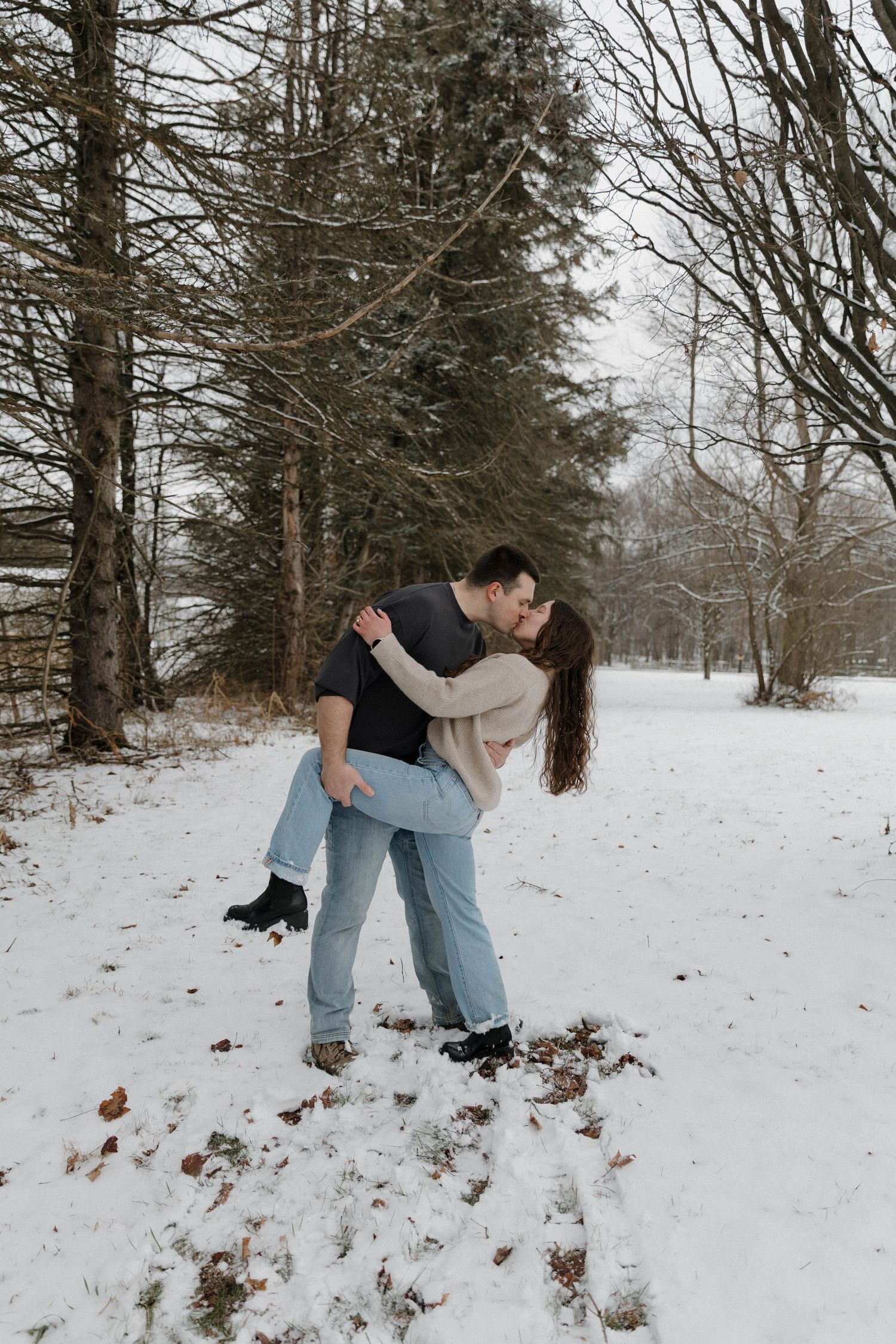 A couple shares a romantic dip pose while standing in a snowy winter landscape surrounded by bare trees.