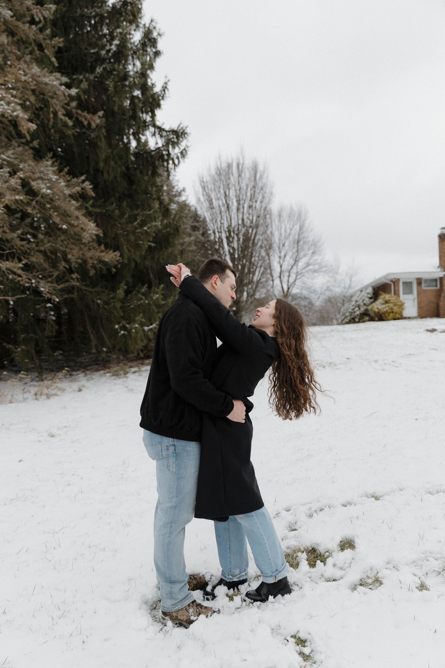 A couple shares romantic moments in a snowy winter landscape with evergreen trees in the background.