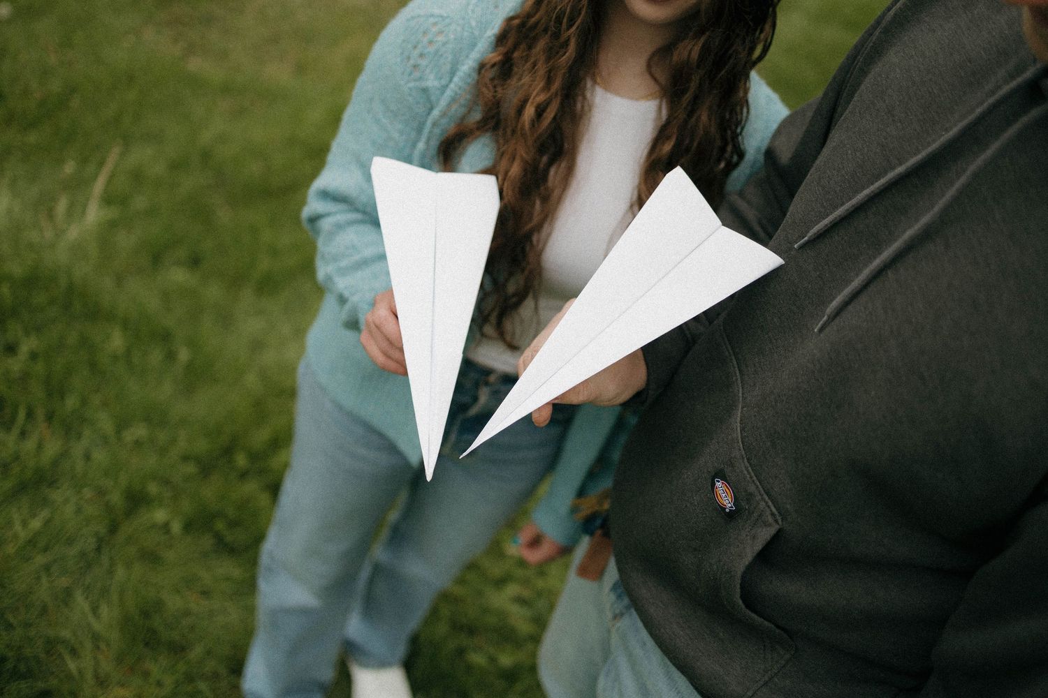 Two people holding white paper airplanes outdoors on green grass, wearing casual blue and gray outfits.