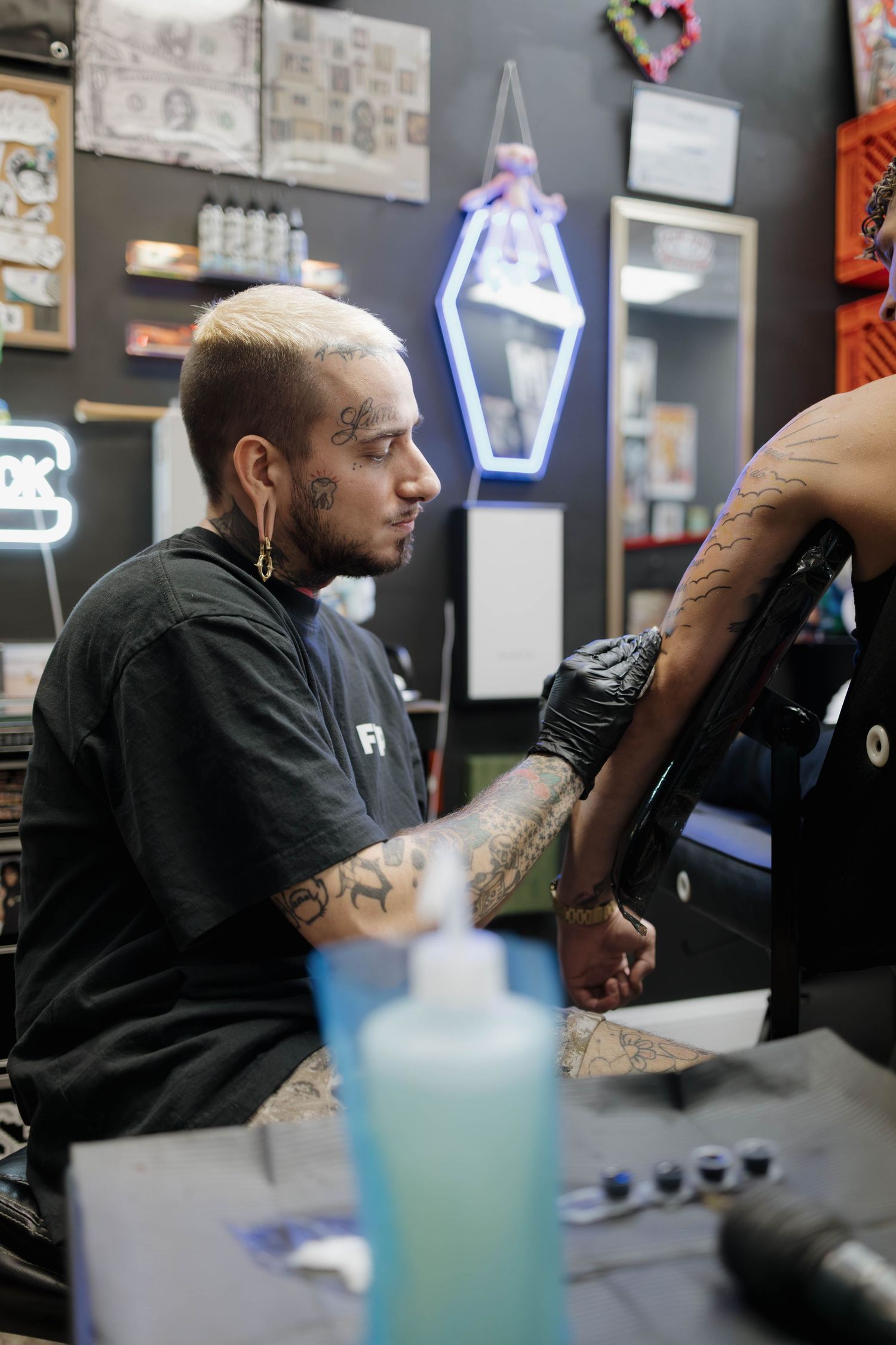 Tattoo artist in black sweater working on a client's arm in a shop with neon signs and artwork on the walls.
