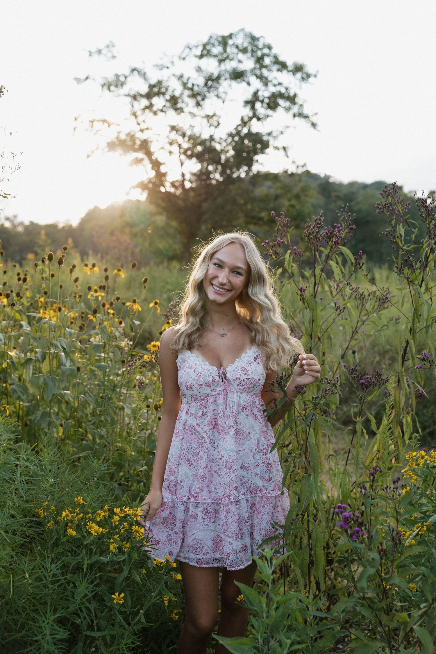 A person in a floral dress smiles while standing in a wildflower field during golden hour.