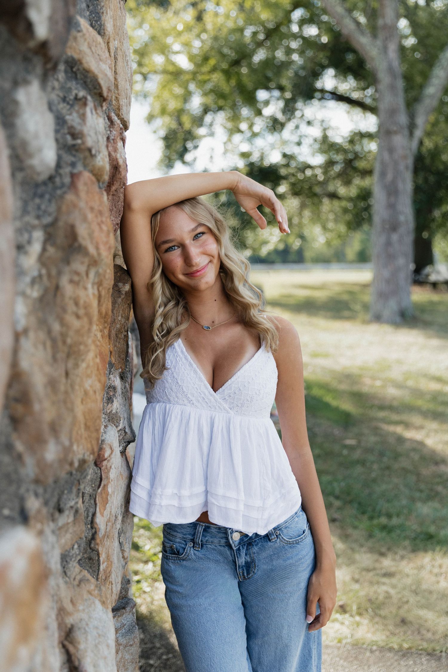 A person in a white sleeveless top and jeans poses casually against tree trunks in a sunny outdoor setting.