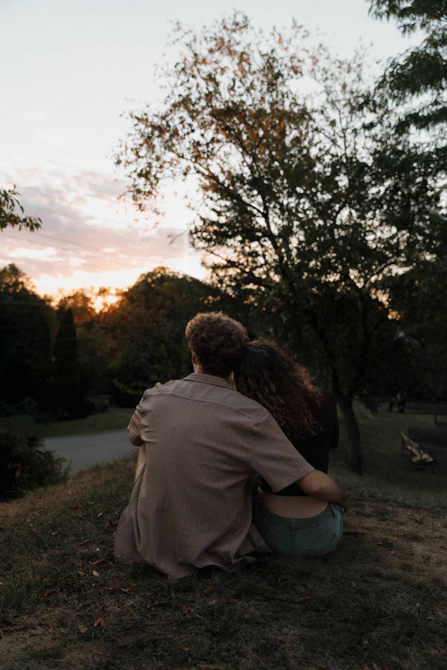 A couple sits together on a hillside at sunset while silhouetted trees frame the peaceful scene in nature.
