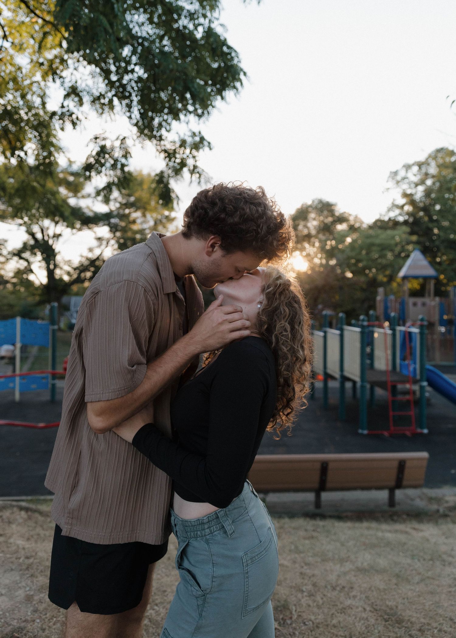 A couple shares a romantic embrace at sunset in a park with playground equipment visible in the background.