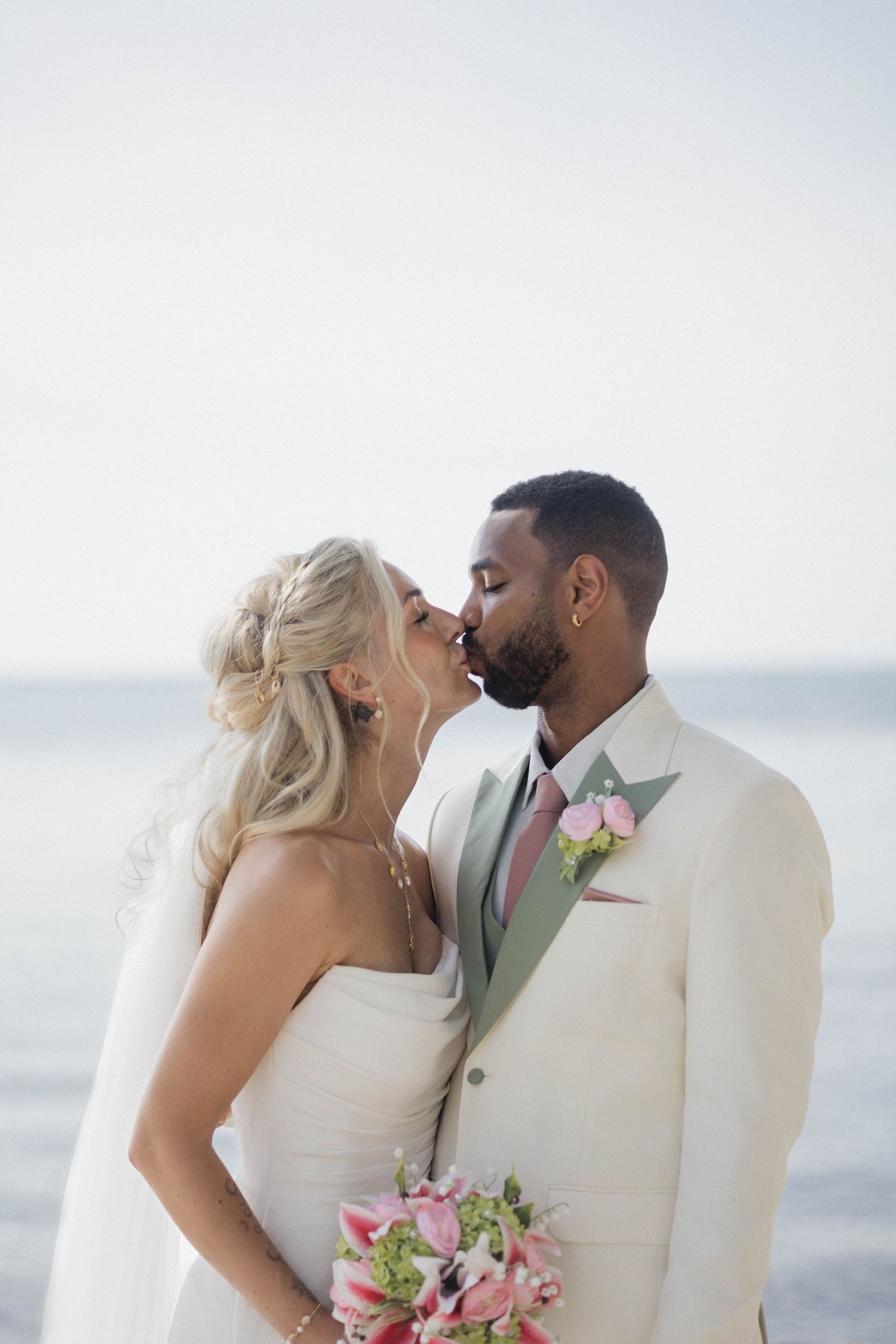 Bride and groom sharing a kiss on the beach, bride holding pink bouquet in white dress, groom in cream suit.