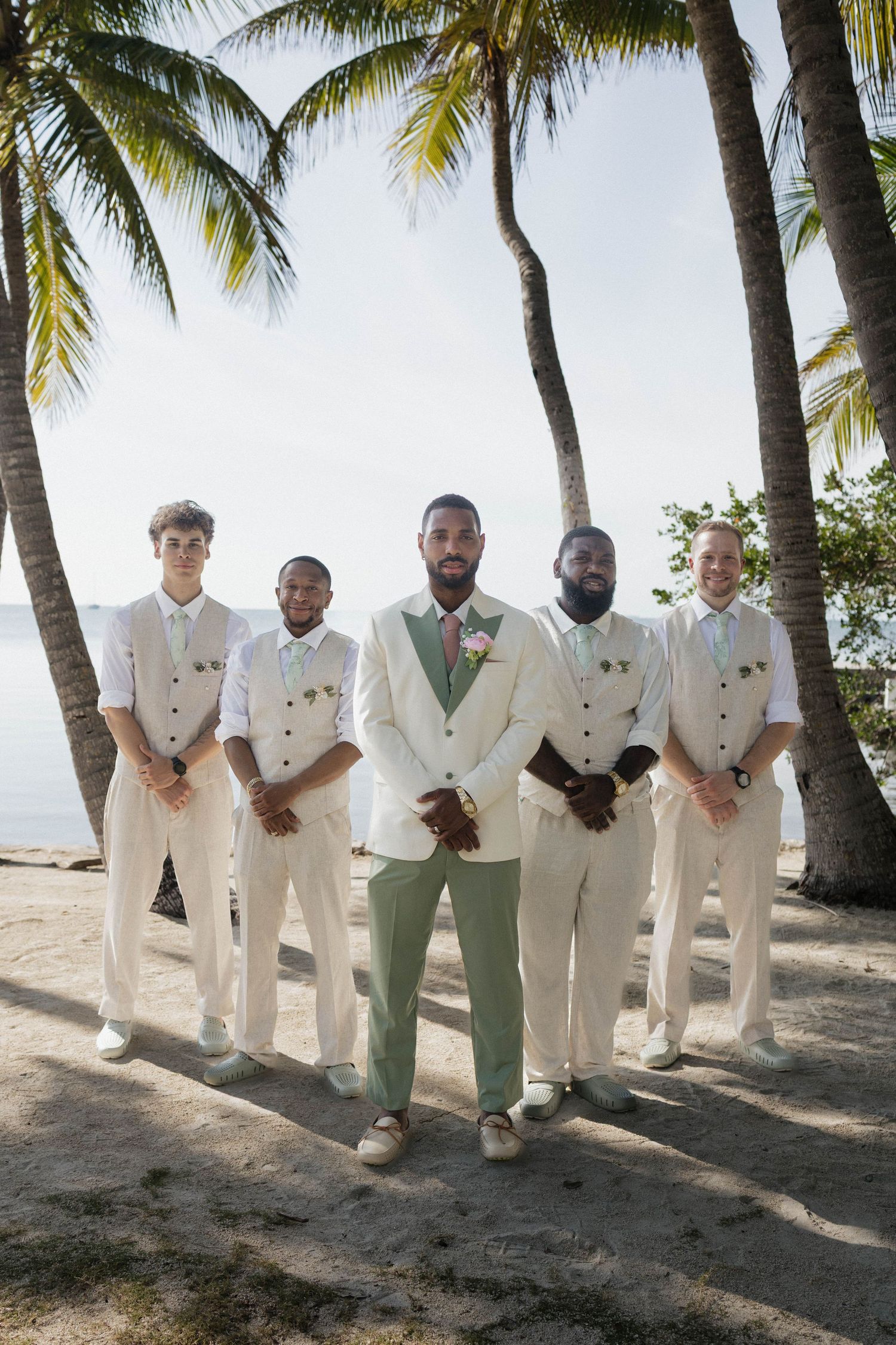 Groom in white suit with groomsmen in matching vests and light pants posing under palm trees on a tropical beach.