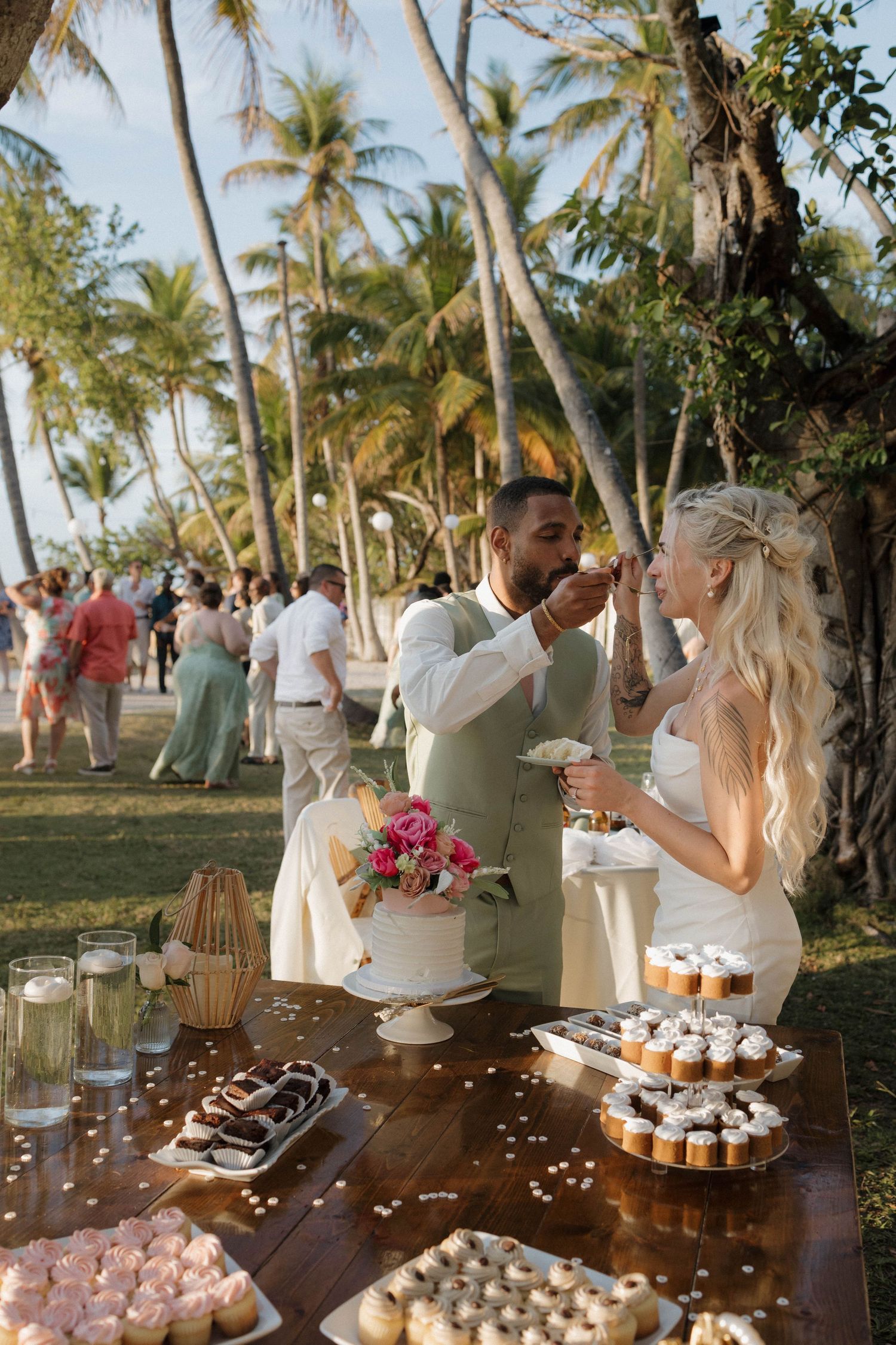Bride and groom feed each other cake at tropical outdoor wedding dessert table with cupcakes and palm trees.