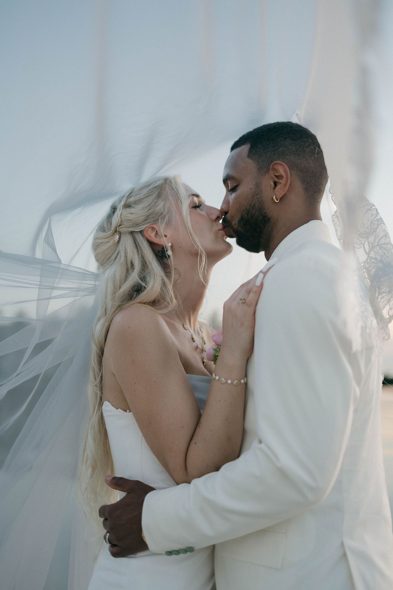 Interracial couple sharing a romantic kiss under a flowing veil, bride in white gown and groom in white suit.