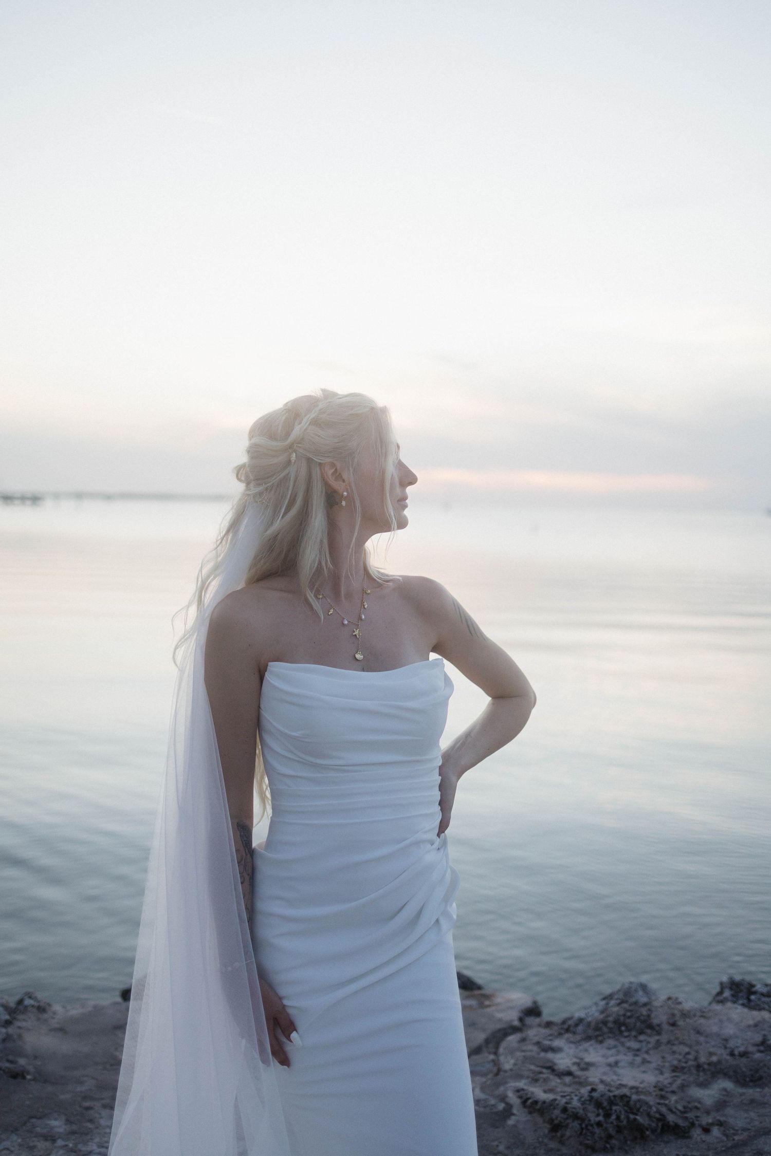 Elegant bride in strapless white gown and veil stands by calm ocean water at sunset.