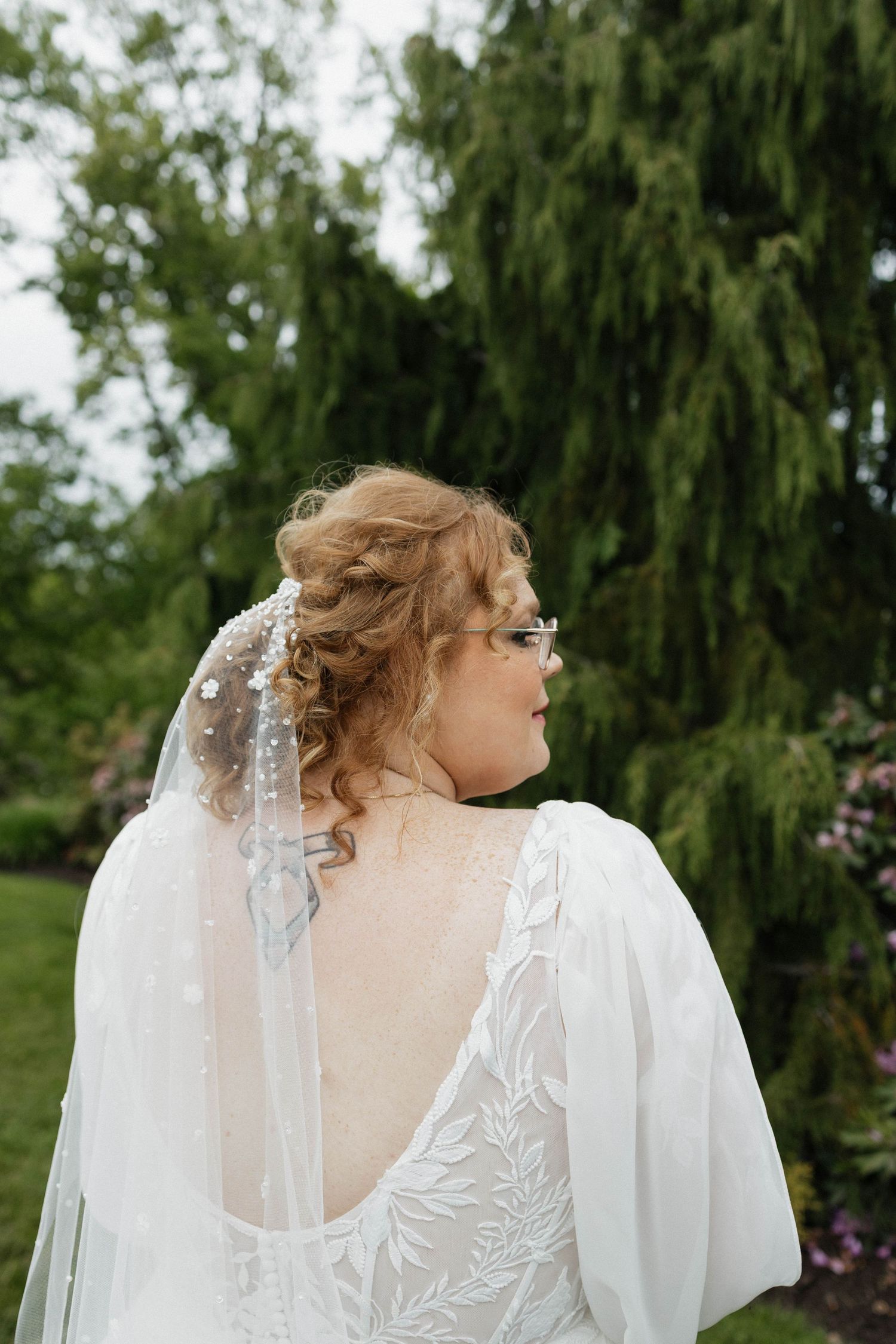 Bride in white lace gown with veil and updo hairstyle stands in lush green garden, viewed from behind.