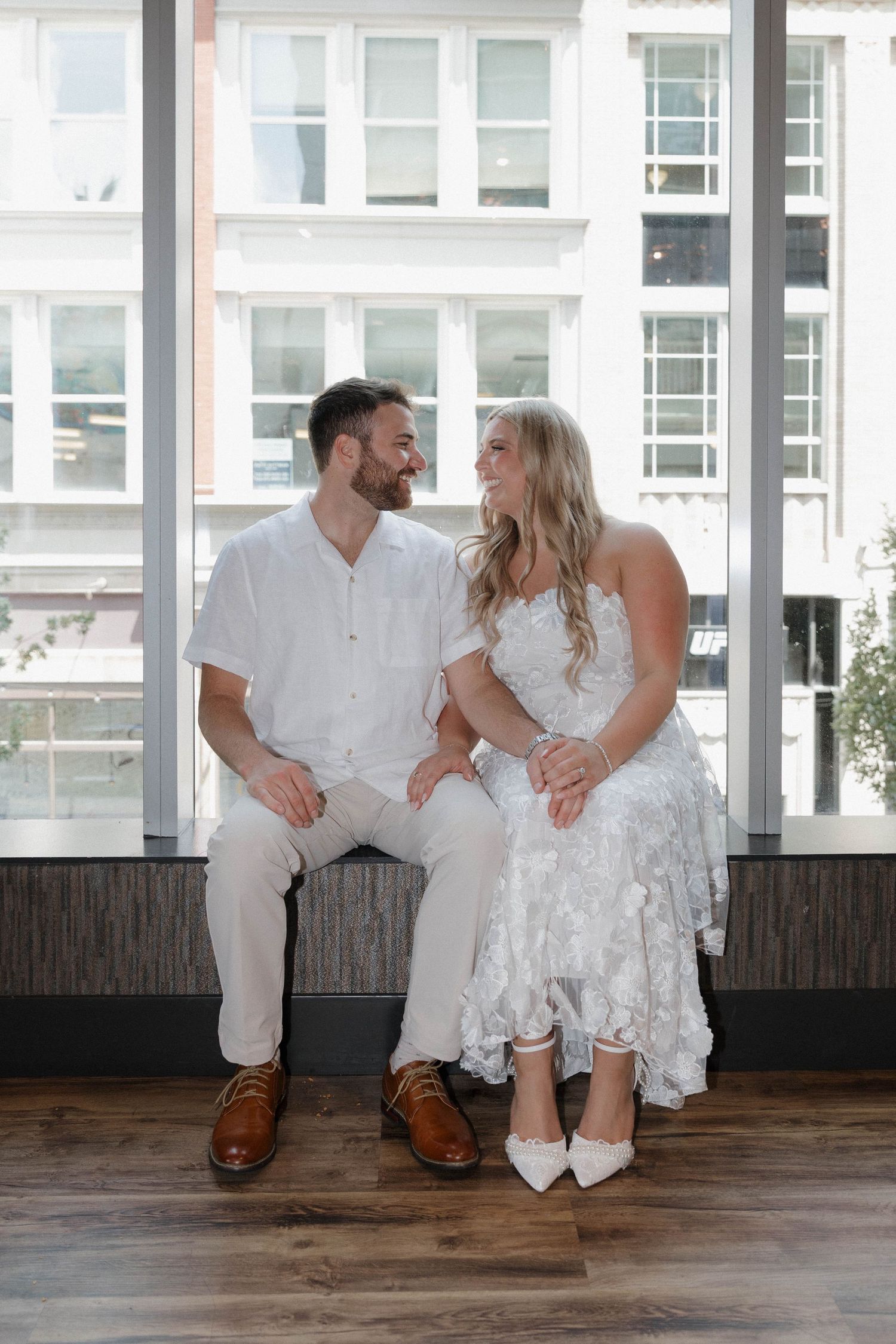 A couple in white wedding attire share a joyful moment while sitting together on a wooden bench by large windows.