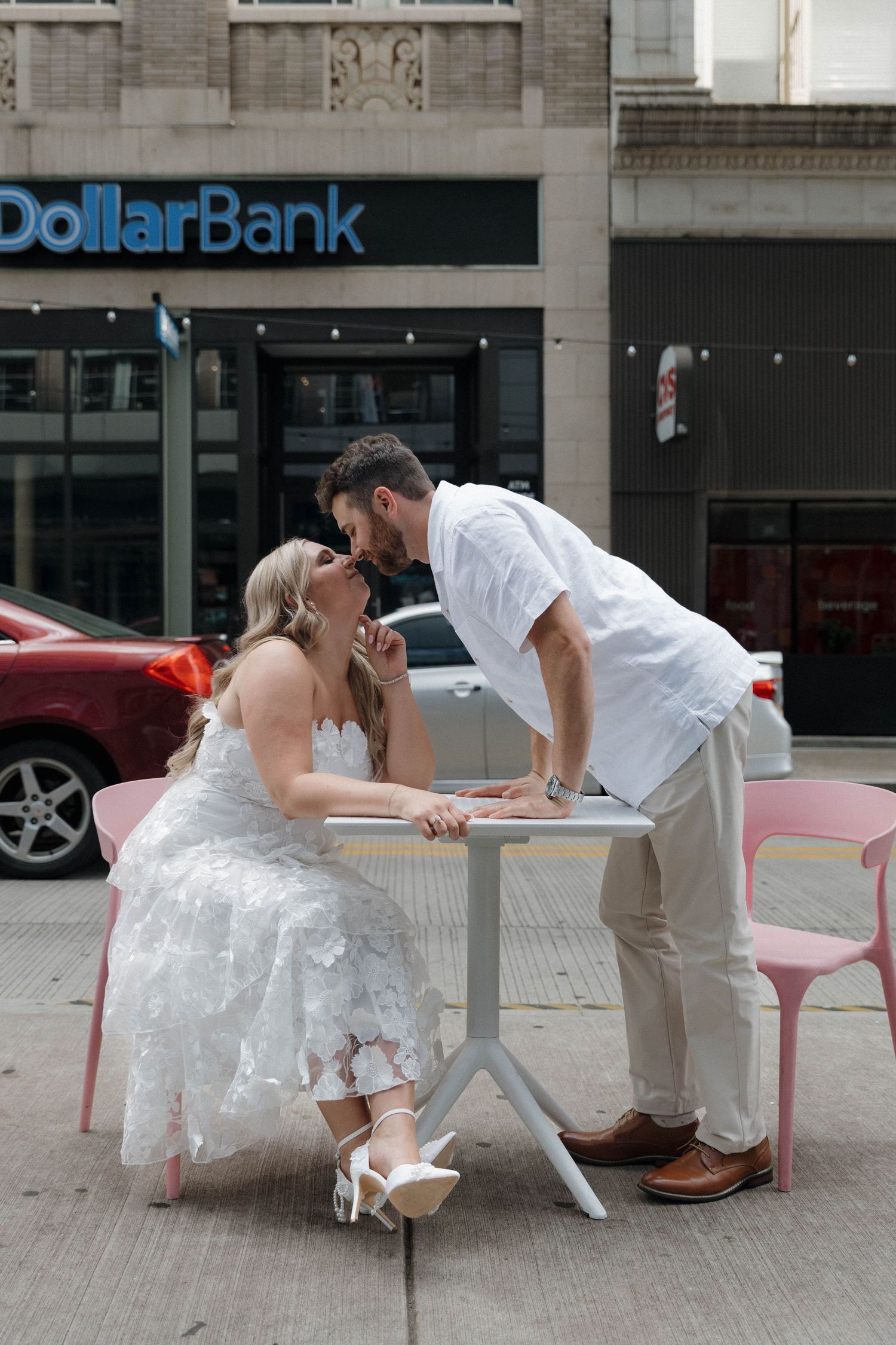 A couple shares a romantic moment at an outdoor cafe table in front of Dollar Bank in an urban setting.