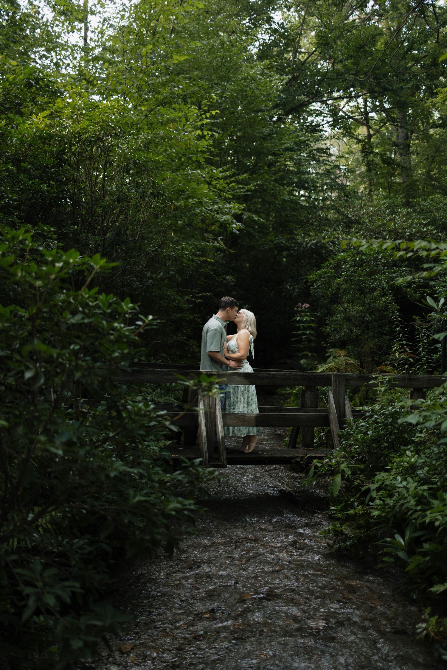 Couple sharing a romantic kiss on a wooden bridge surrounded by lush green forest trees.