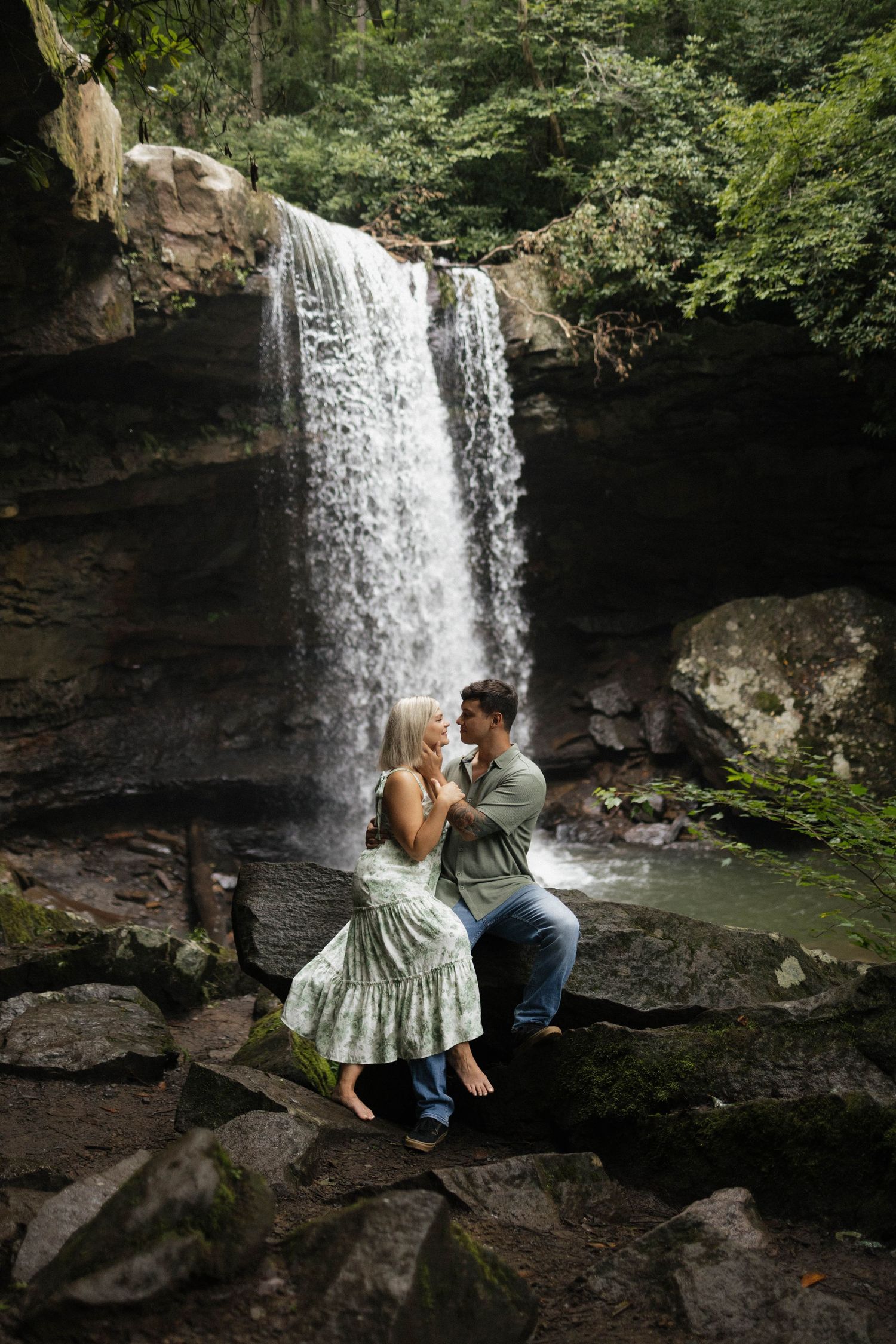 Couple sitting on rocks in front of a stunning waterfall surrounded by lush green forest.