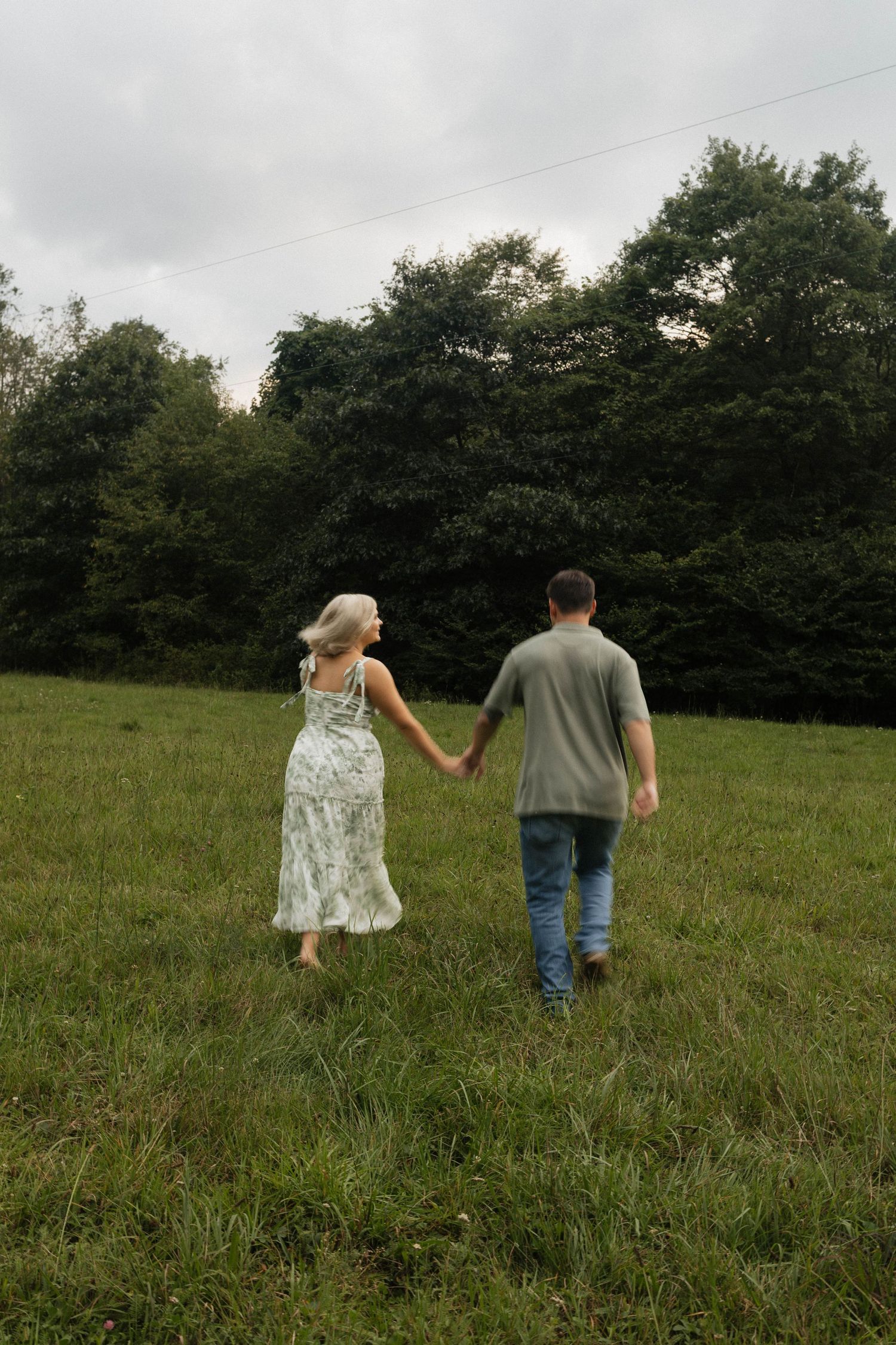 A couple holds hands walking through a lush green meadow toward a dense tree line under an overcast sky.