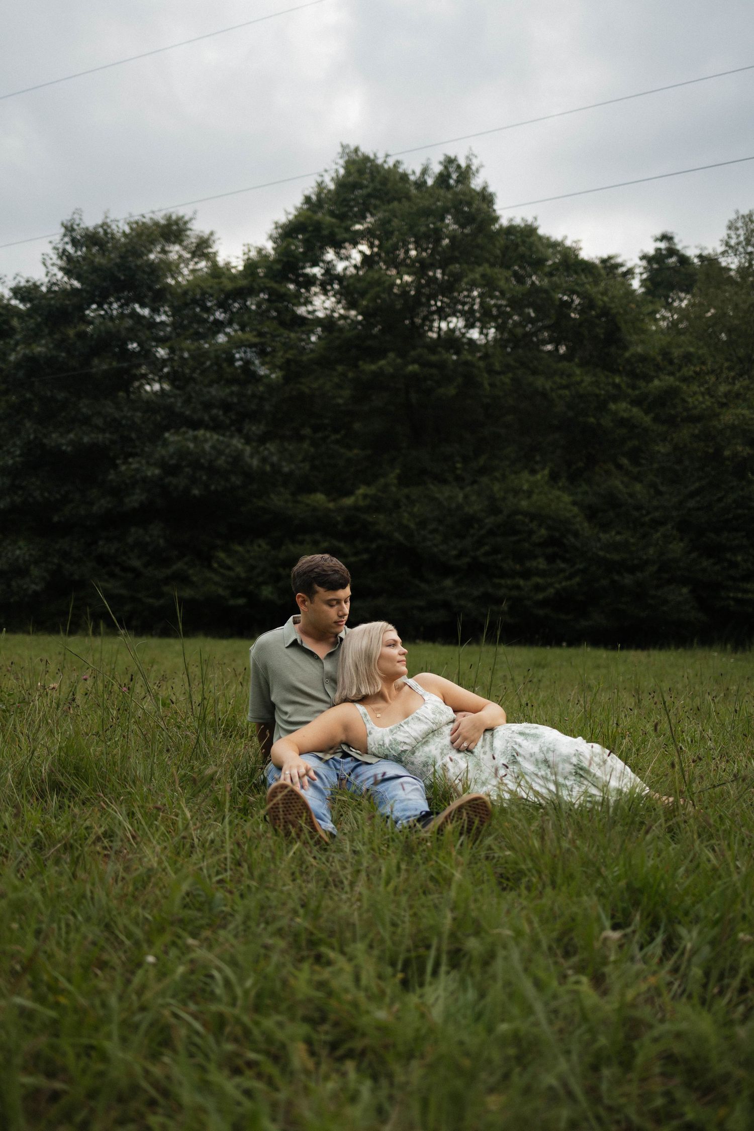 Couple sitting together in a lush green field, woman in floral dress leaning on man in gray shirt, trees behind.
