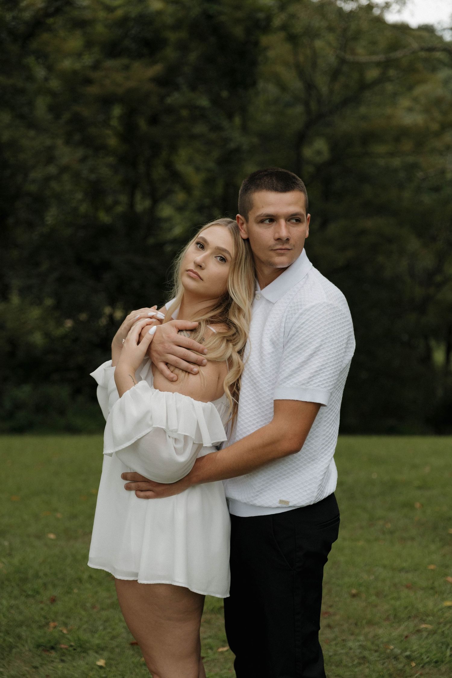 A couple in white clothing embraces tenderly in a green outdoor setting during a romantic photoshoot.