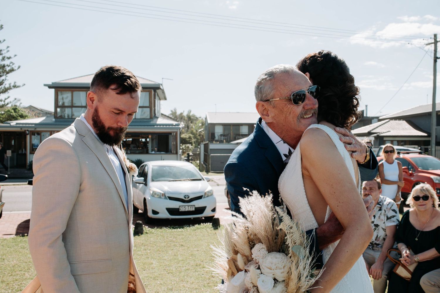 Beachside wedding ceremony with wooden arch and guests seated on grass.