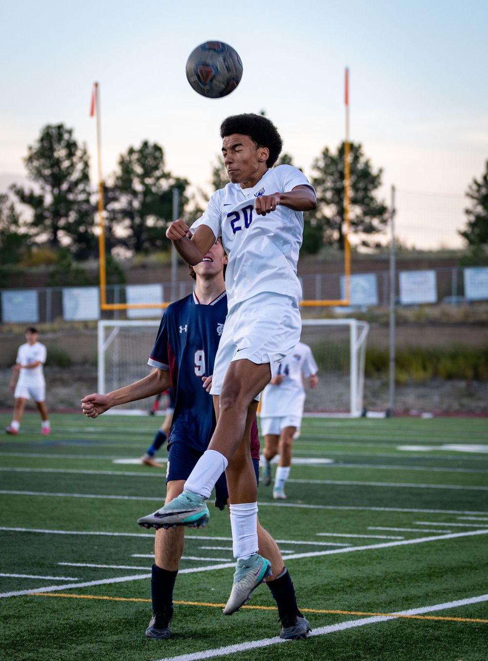 Soccer player in white jersey jumps high to head the ball during competitive match.