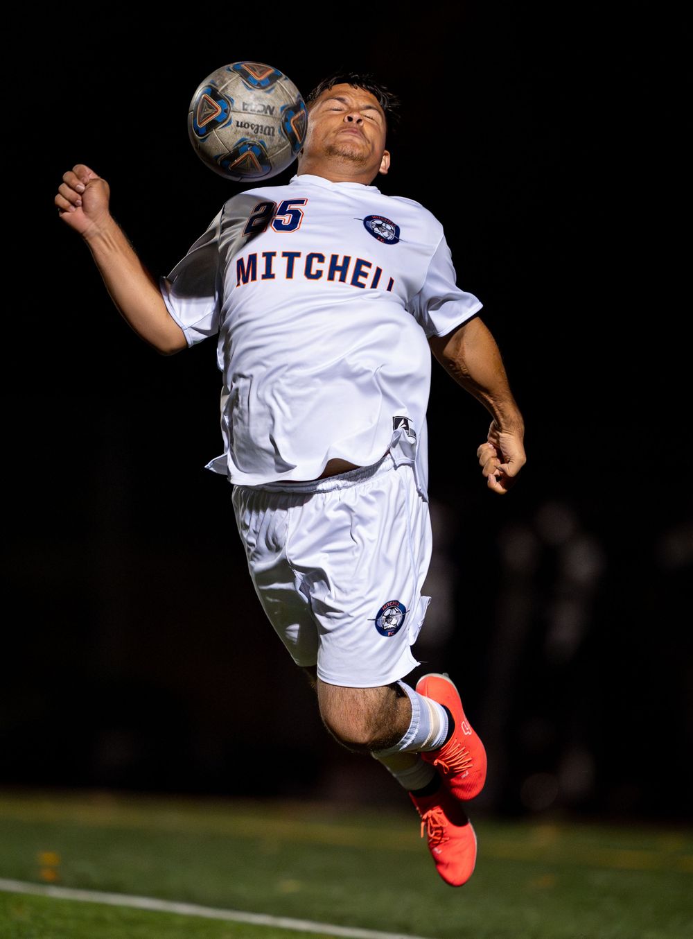 Soccer player in white Mitchell uniform jumps to control ball against dark background.