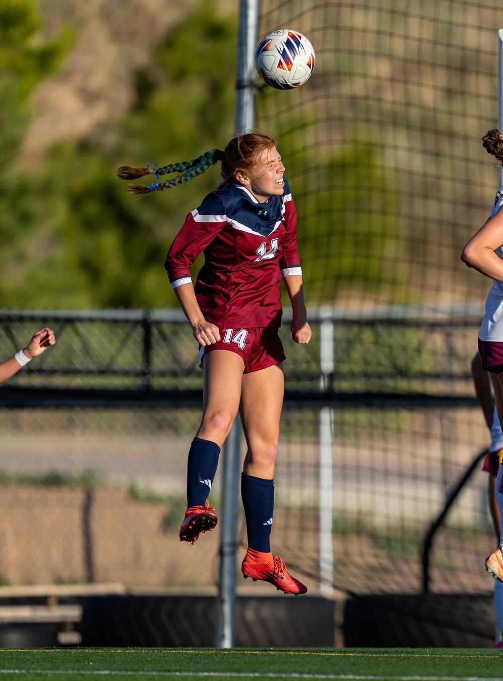 Soccer player wearing maroon jersey jumps high to head the ball during competitive match on outdoor field.