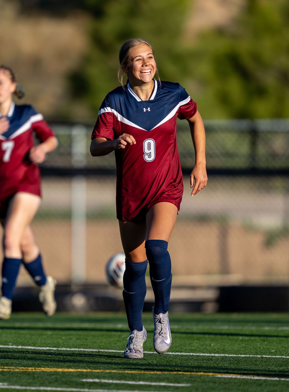 A female soccer player wearing a red jersey with number 9 runs on an artificial turf field during a match.