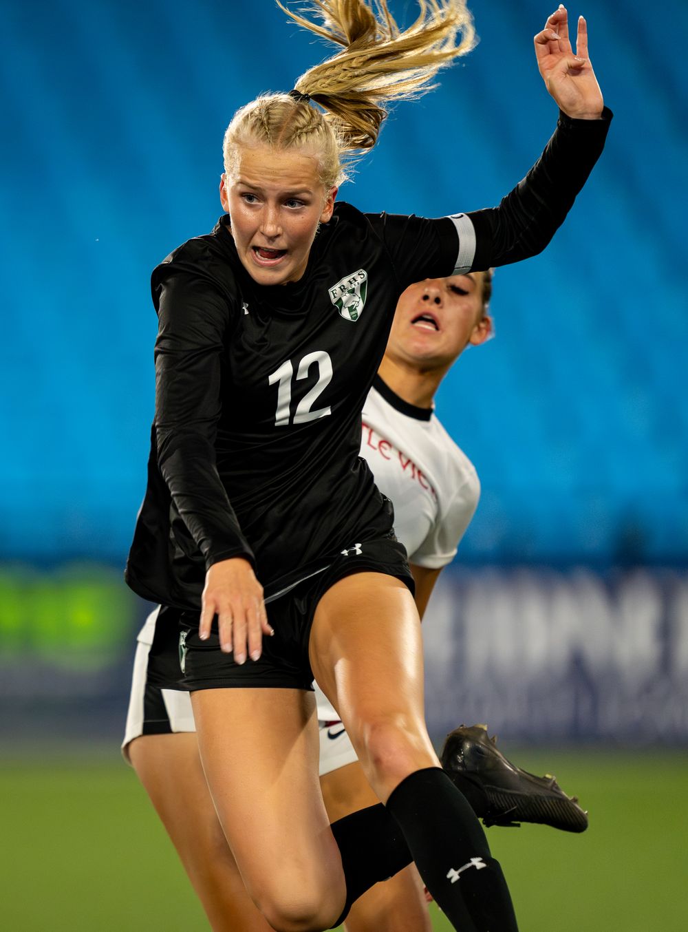 Two soccer players in a competitive match leap high in the air, one in black jersey number 12 and another in white jersey.