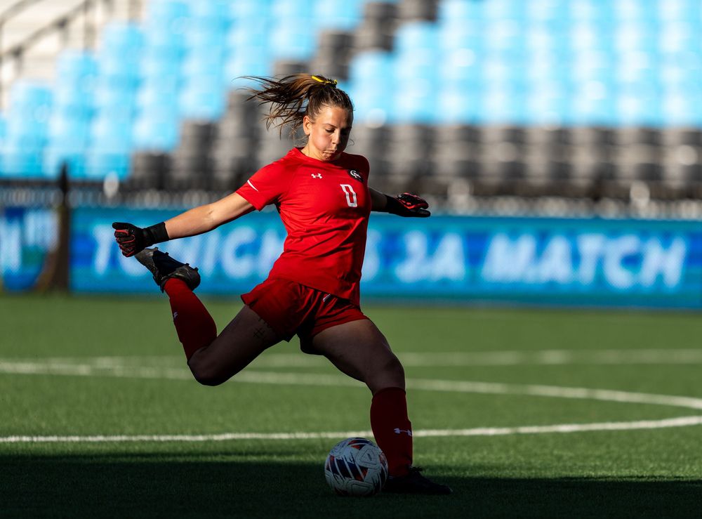 Athletic soccer player in red uniform performs high kick during match in stadium with blue seats.