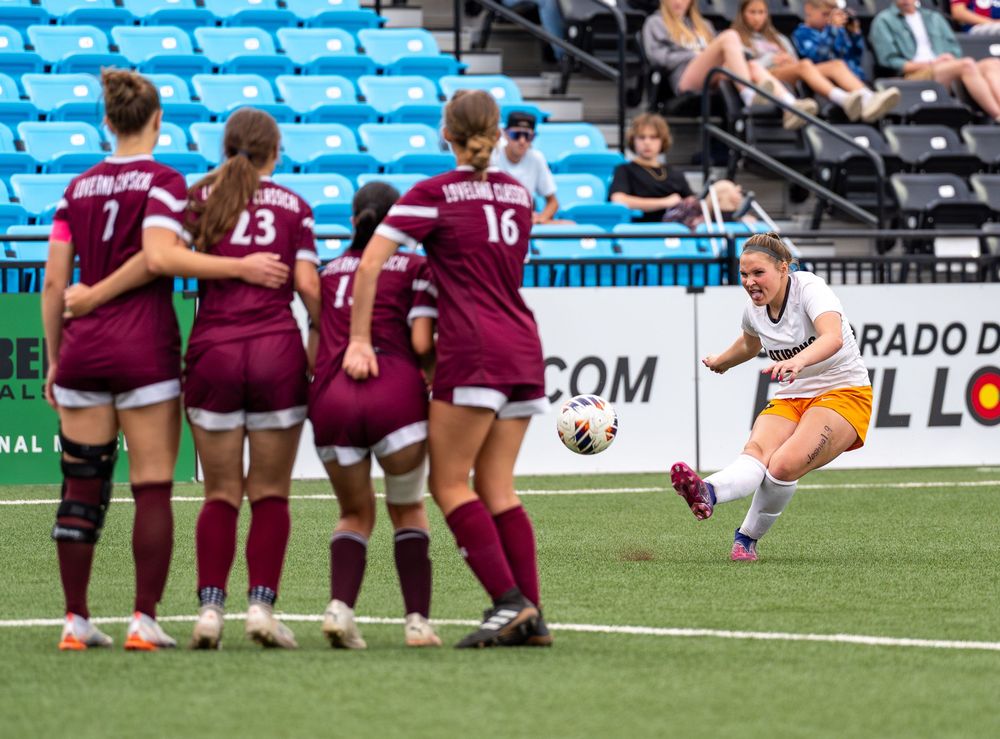 Soccer player in orange and white jumps past defensive wall wearing burgundy uniforms during match at stadium.
