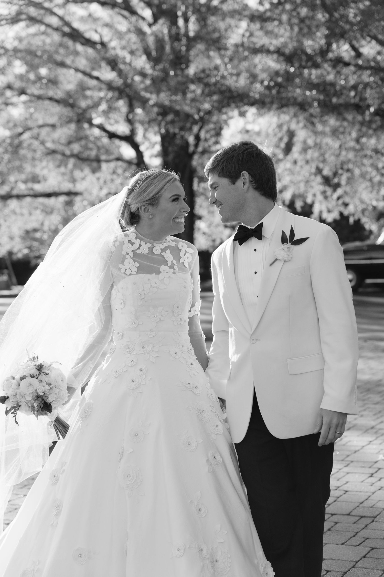 Newlywed couple shares a romantic moment outdoors in black and white, with bride in lace gown and groom in white jacket.