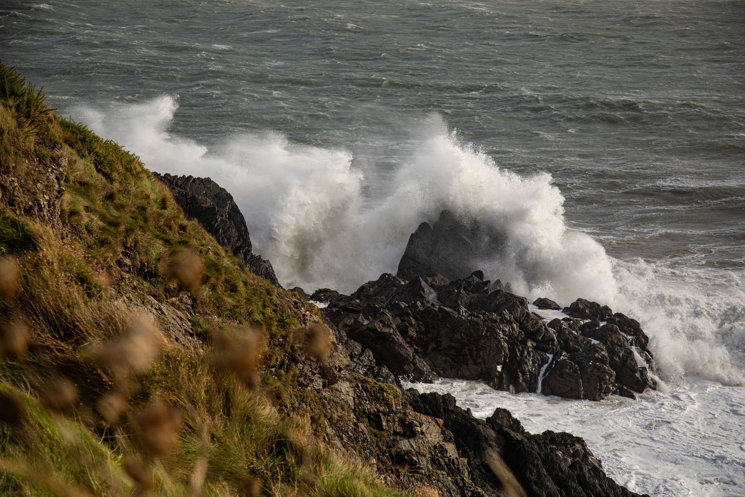 Powerful ocean waves crash against rocky coastal cliffs as stormy seas create dramatic white spray in choppy conditions.