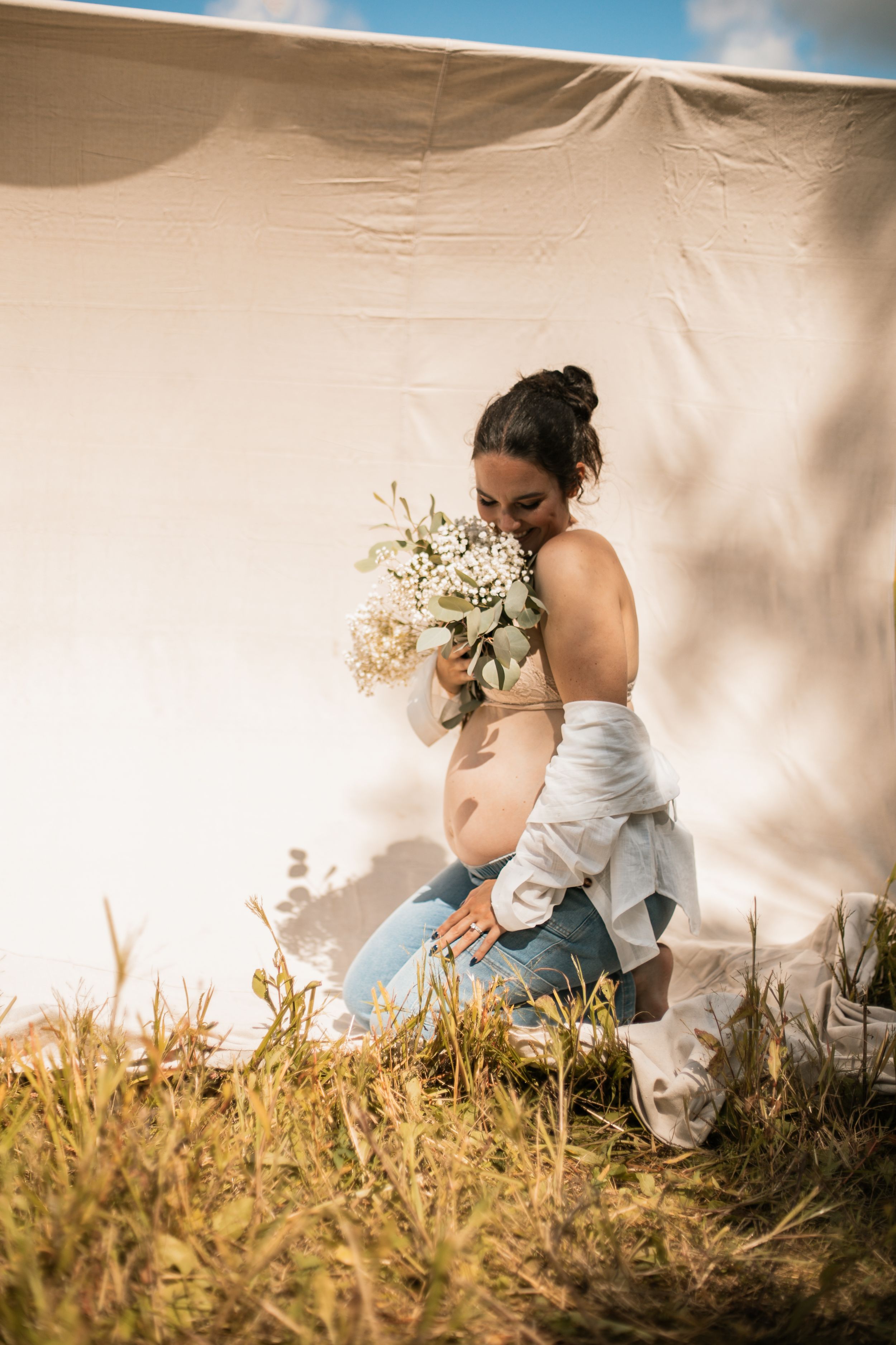 A scenic outdoor photo showing someone in a white dress kneeling in tall grass while holding a bouquet of flowers.