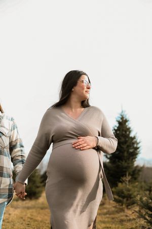 Expectant woman in beige wrap dress walks outdoors while holding hands with partner during maternity photoshoot.