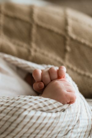 A tiny baby foot with delicate toes peeks out from under a white knitted blanket against a beige background.