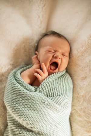 A yawning infant wrapped in a mint green knitted blanket lies on a cream-colored plush surface.