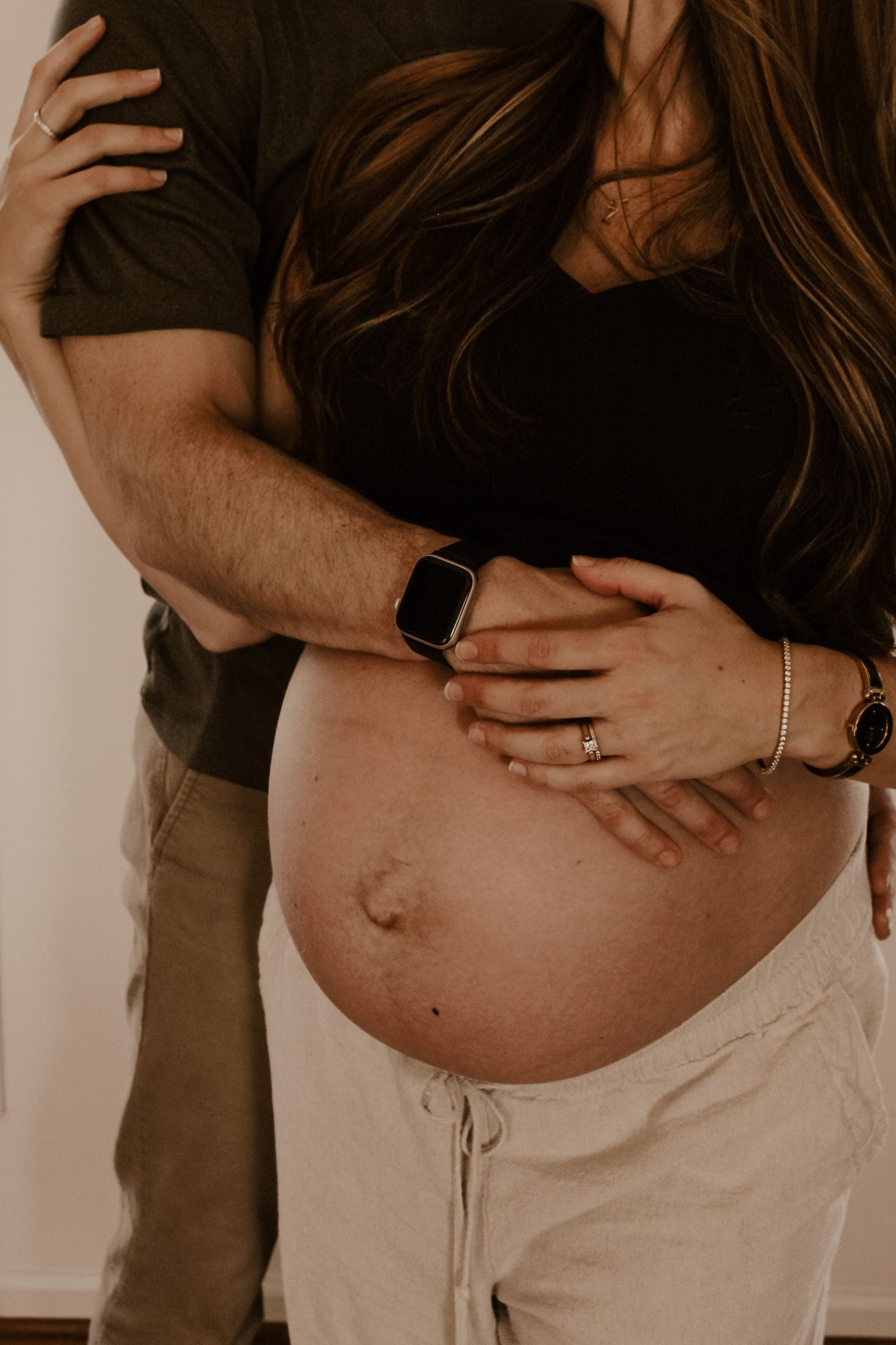 Intimate pregnancy photo showing hands resting on a bare pregnant belly with an Apple watch and jewelry visible.