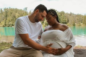 Intimate maternity photo of expecting couple embracing by turquoise lake with forest backdrop.