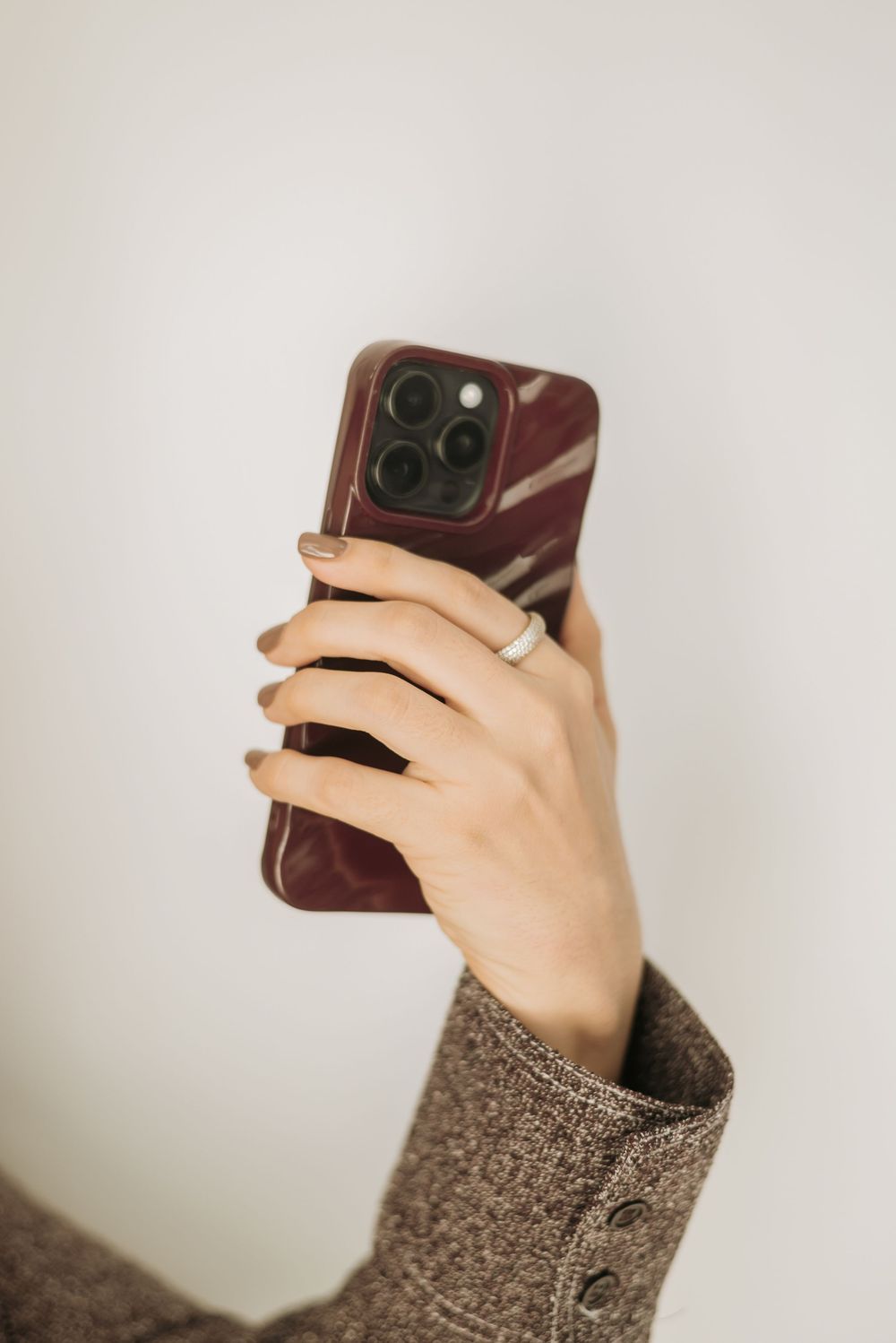 A hand wearing a brown sweater sleeve holds up a burgundy iPhone 13 Pro against a light grey background.