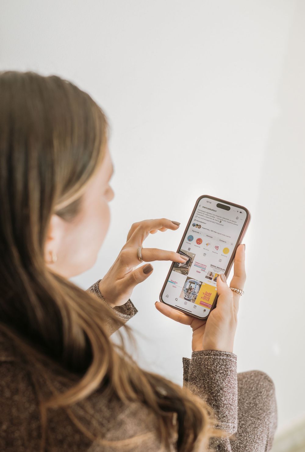 Person in gray sweater browsing social media on smartphone against white wall.