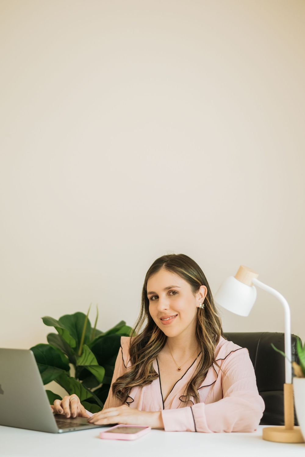 A professional sits at a desk with laptop, white table lamp, and houseplant in a bright home office setting.