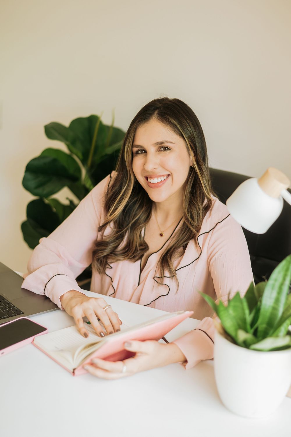 Someone wearing pink sits at desk with houseplants, writing in notebook while smiling in bright workspace.