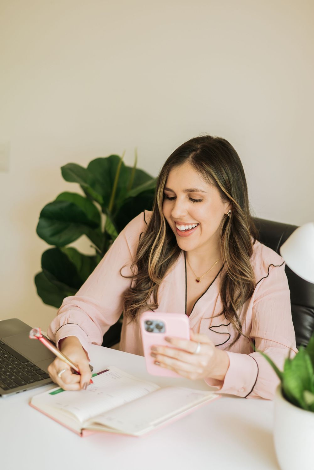 Woman in pink blouse smiles while working at desk with notebook and phone near houseplant.